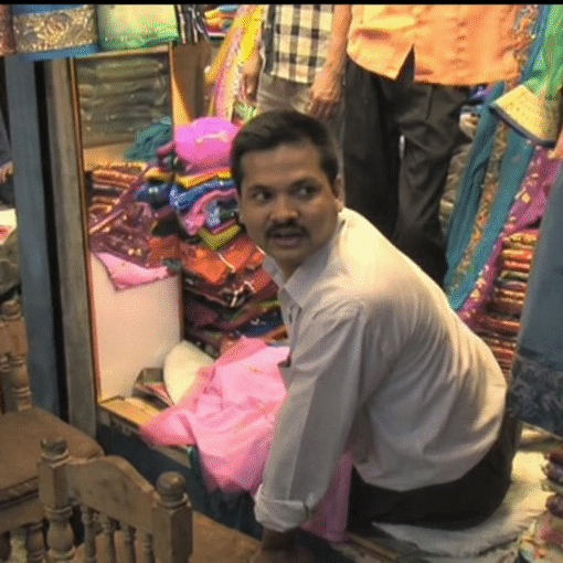 Hombre bangladesí agachado en una tienda de telas, rodeado de vestidos y tejidos de colores, mirando hacia un lado mientras ordena mercancía en un espacio comercial estrecho.