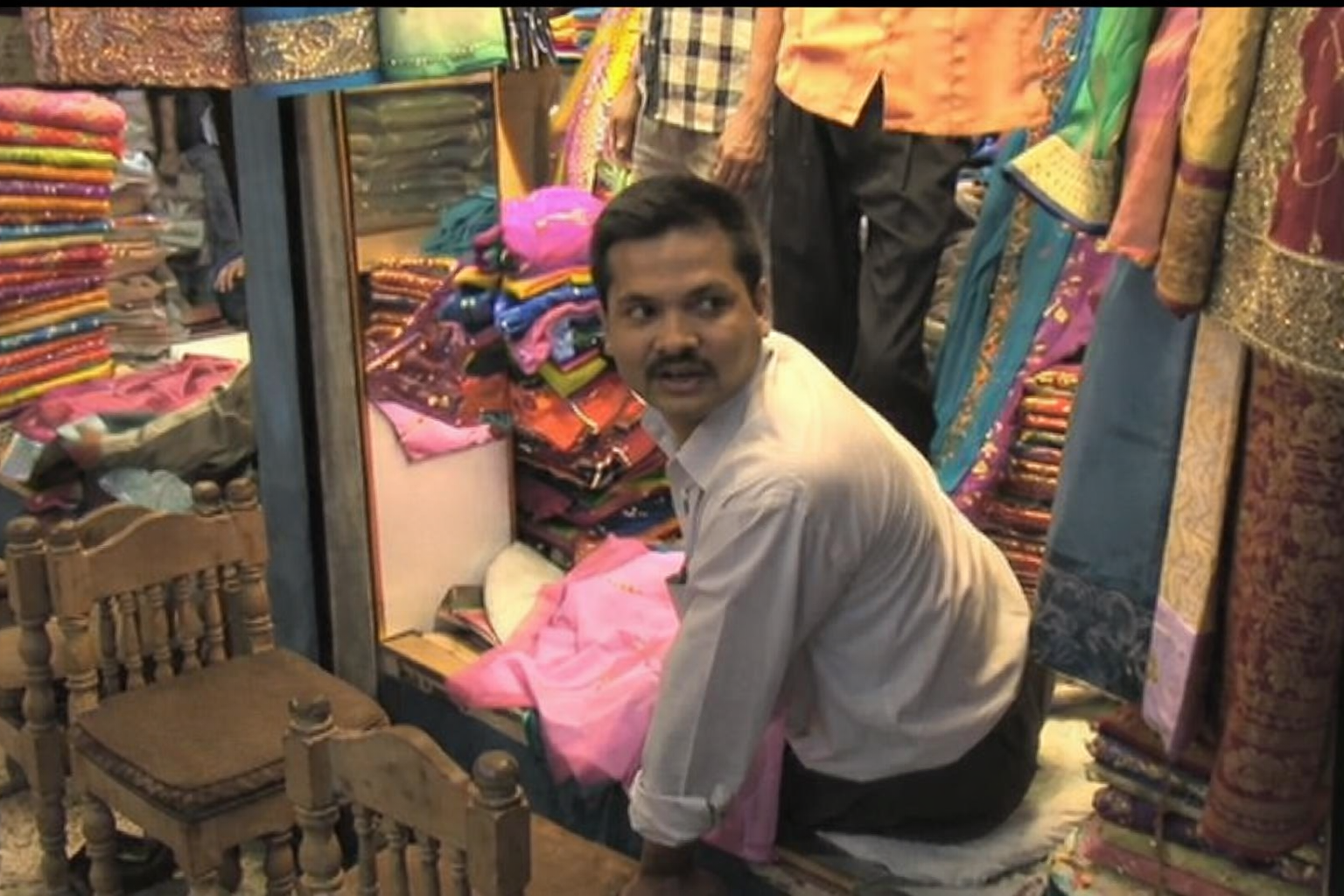 Hombre bangladesí agachado en una tienda de telas, rodeado de vestidos y tejidos de colores, mirando hacia un lado mientras ordena mercancía en un espacio comercial estrecho.