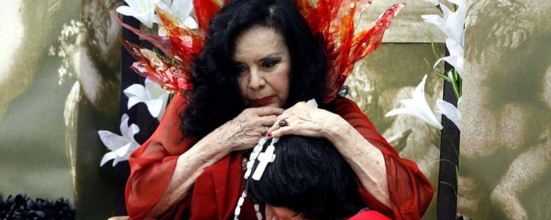 Una mujer con vestido rojo y pelo negro está sentada y mira hacia el suelo.