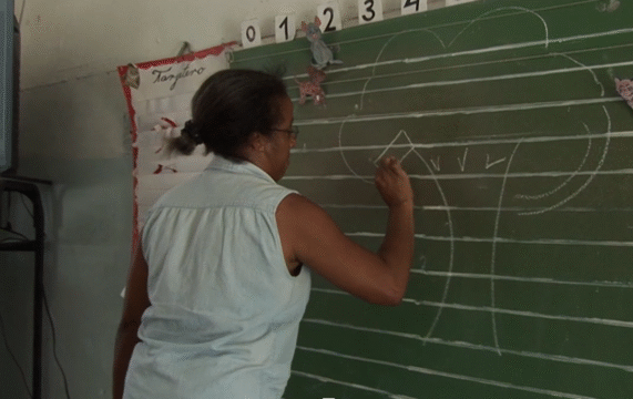 Profesora de espalda a la clase, dibujando un árbol con tiza sobre la pizarra. En la pizarra se pueden ver varios dibujos de animales que parecen coloreados por niños. La profesora viste una chaqueta vaquera sin mangas y lleva coleta y gafas.