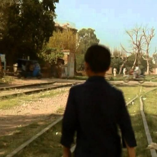 La imagen muestra a un niño caminando por unas vías de tren, visto desde atrás. El niño está de espaldas en el centro de la escena.