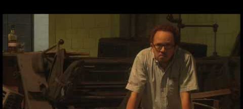 Fotograma de "La bodega". Hombre sentado entre barullo de objetos rotos y viejos con las manos en las rodillas.