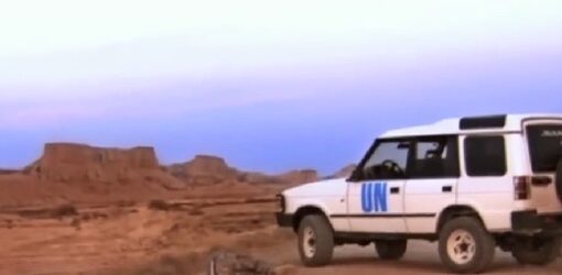 Un vehículo blanco con el emblema de la ONU (UN) situado en un paisaje árido y desértico, rodeado de formaciones rocosas