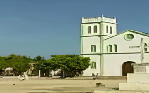 Fotograma del documental Viajero de mi mismo. Hay una iglesia de color blanco al fondo, es un día soleado sin nubes. esta en medio de una plaza y hay arboles y personas andando.