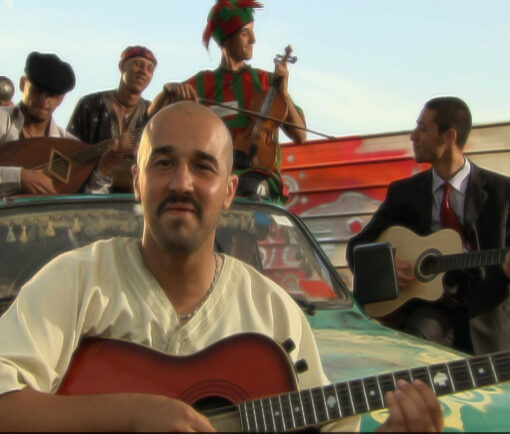 La plaza. Fotograma del largometraje. Muestra a un hombre encima de un coche con una guitarra, mirando a cámara, apoyado en un coche donde hay más músicos.