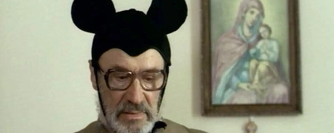 Niki y Flo. Fotograma del largometraje. Se ve a un hombre mayor que a pesar de su porte serio, tiene puesto un gorro de Mickey Mouse.