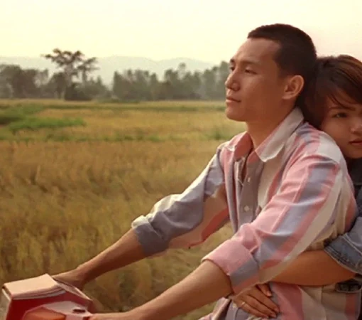 Escena de la película Terriblemente Alegre. Una pareja joven viaja en una motocicleta por un camino rural rodeado de campos. El hombre conduce mirando hacia adelante con expresión pensativa, mientras la mujer, sentada detrás de él, lo abraza por la cintura y apoya la cabeza en su espalda. Al fondo se ven campos abiertos y montañas suaves bajo una luz cálida de atardecer.