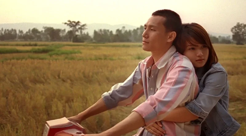 Escena de la película Terriblemente Alegre. Una pareja joven viaja en una motocicleta por un camino rural rodeado de campos. El hombre conduce mirando hacia adelante con expresión pensativa, mientras la mujer, sentada detrás de él, lo abraza por la cintura y apoya la cabeza en su espalda. Al fondo se ven campos abiertos y montañas suaves bajo una luz cálida de atardecer.