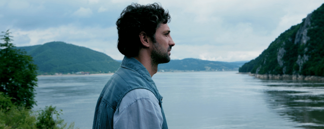 Hombre de pelo castaño y chaqueta vaquera mirando hacia el horizonte con un lago de fondo.