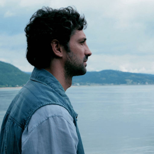 Hombre de pelo castaño y chaqueta vaquera mirando hacia el horizonte con un lago de fondo.