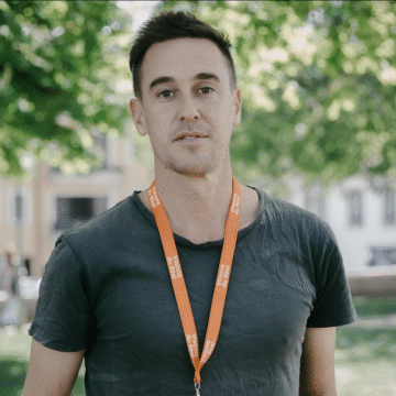 Retrato de un hombre joven al aire libre con camiseta oscura y una acreditación naranja colgada al cuello, con árboles y edificios desenfocados de fondo.