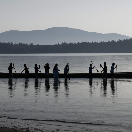 Una canoa larga se encuentra parcialmente en el agua, con once personas en pie a lo largo de su costado, sosteniendo remos.