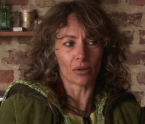 Una mujer mayor con un sueter verde oscuro y una camisa verde manzana que dirige su vista hacia un lado mientras habla tiene pelo corto