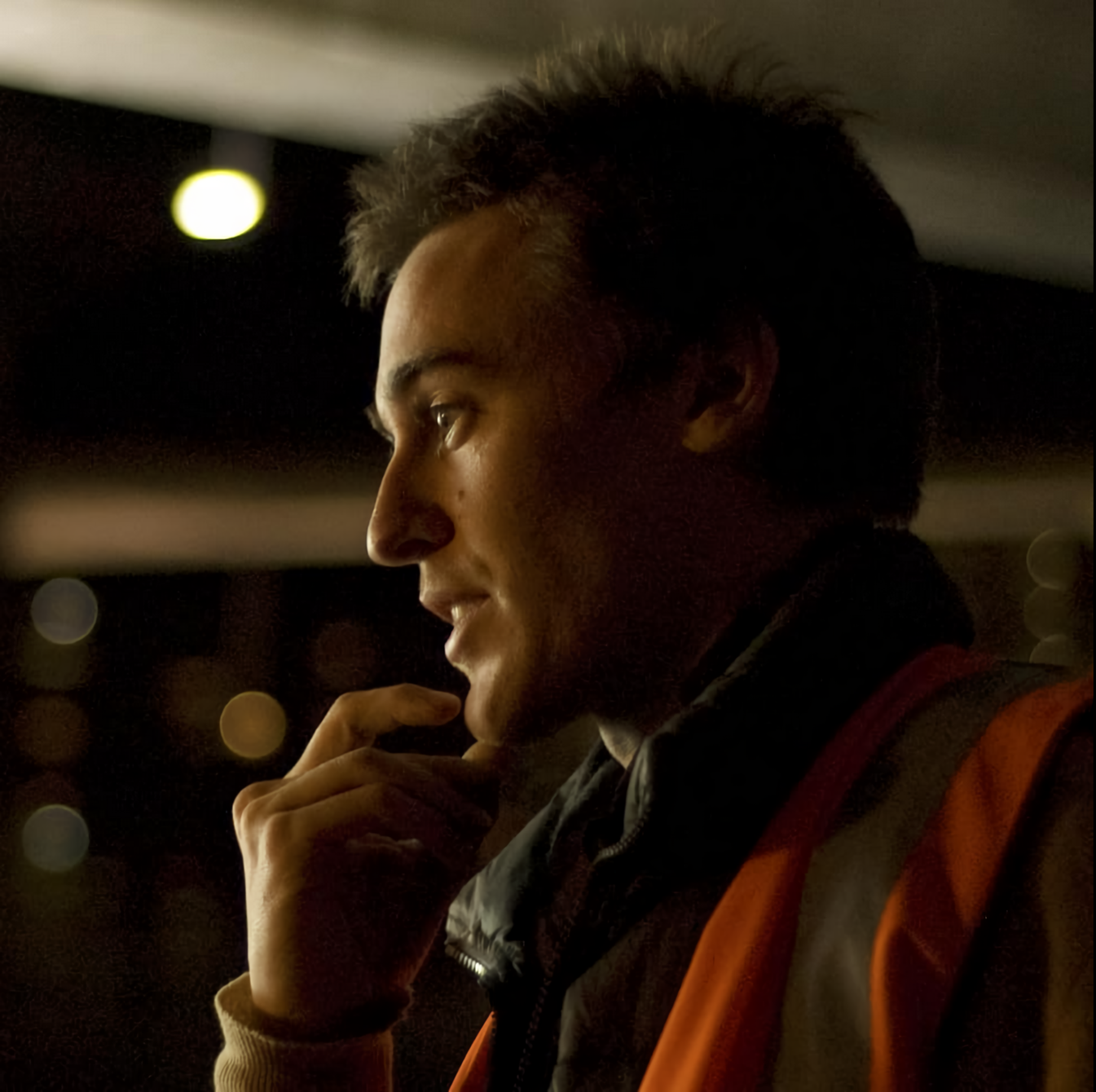 Retrato lateral de un hombre joven con chaleco reflectante naranja, pensativo, iluminado por luces nocturnas.