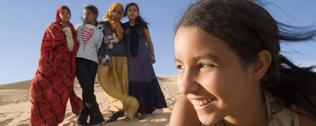 Niña saharaui sonriente en primer plano con cuatro mujeres al fondo en un paisaje desértico.