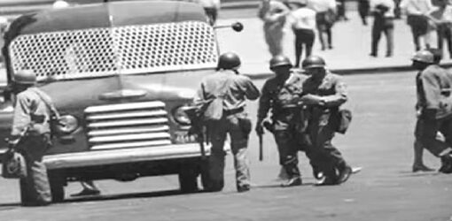 Un grupo de soldados armados rodea un camión con una rejilla protectora en el parabrisas. Al fondo, varias personas observan la escena desde la acera.