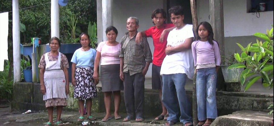Siete personas, entre ellas hombres, mujeres y una niña, están de pie frente a una casa con columnas desgastadas. Posan en fila sobre un suelo húmedo, con expresiones serias y un entorno verde y tropical detrás.
