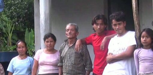 Siete personas, entre ellas hombres, mujeres y una niña, están de pie frente a una casa con columnas desgastadas. Posan en fila sobre un suelo húmedo, con expresiones serias y un entorno verde y tropical detrás.