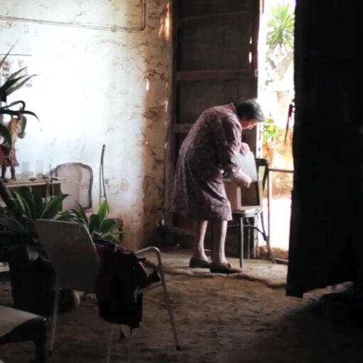 Una anciana, vestida con una bata de flores, se inclina con delicadeza sobre una silla junto a la puerta abierta de una estancia austera. La luz que entra suavemente realza la calma y la melancolía del momento.