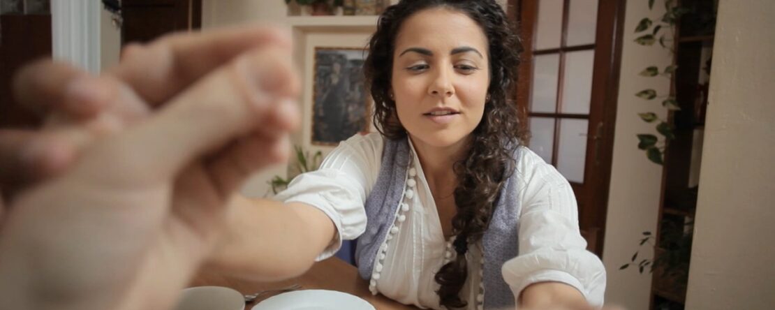 Una mujer joven de cabello oscuro y rizado, con blusa blanca y chaleco gris, sentada a una mesa de madera interactuando con alguien fuera de cámara.