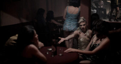 Mariposas carmesí. Fotograma del corto. La imagen presenta el interior de un bar o club nocturno, con un ambiente acogedor y tenue iluminación. Sentadas alrededor de esta mesa hay dos personas.