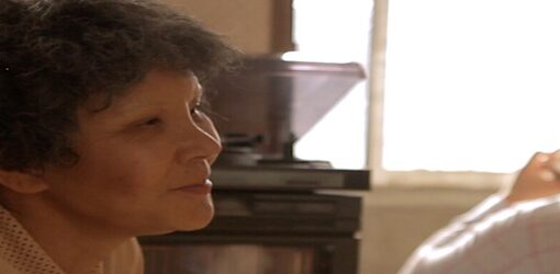 Una mujer mayor, de perfil y con gesto sereno, conversa en una habitación iluminada por la luz de una ventana, mientras otra persona permanece de espaldas al fondo.