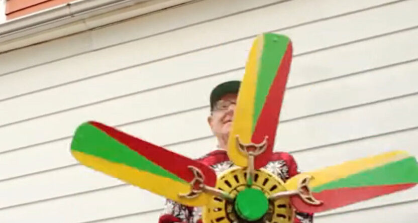 En primer plano, un hombre sostiene una hélice decorada por él con colores rojo, verde y amarillo.