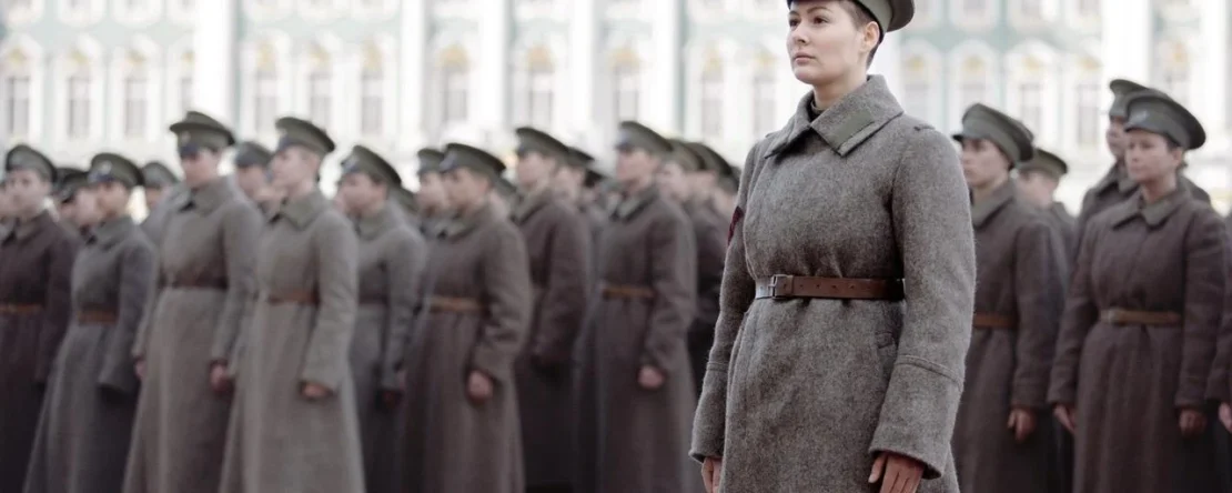 Fotograma de "Batallón" en el que aparece una mujer vestida de militar con un abrigo gris y una gorra. Detrás, hay todo un ejército de personas vestidas como ella.