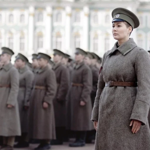 Fotograma de "Batallón" en el que aparece una mujer vestida de militar con un abrigo gris y una gorra. Detrás, hay todo un ejército de personas vestidas como ella.