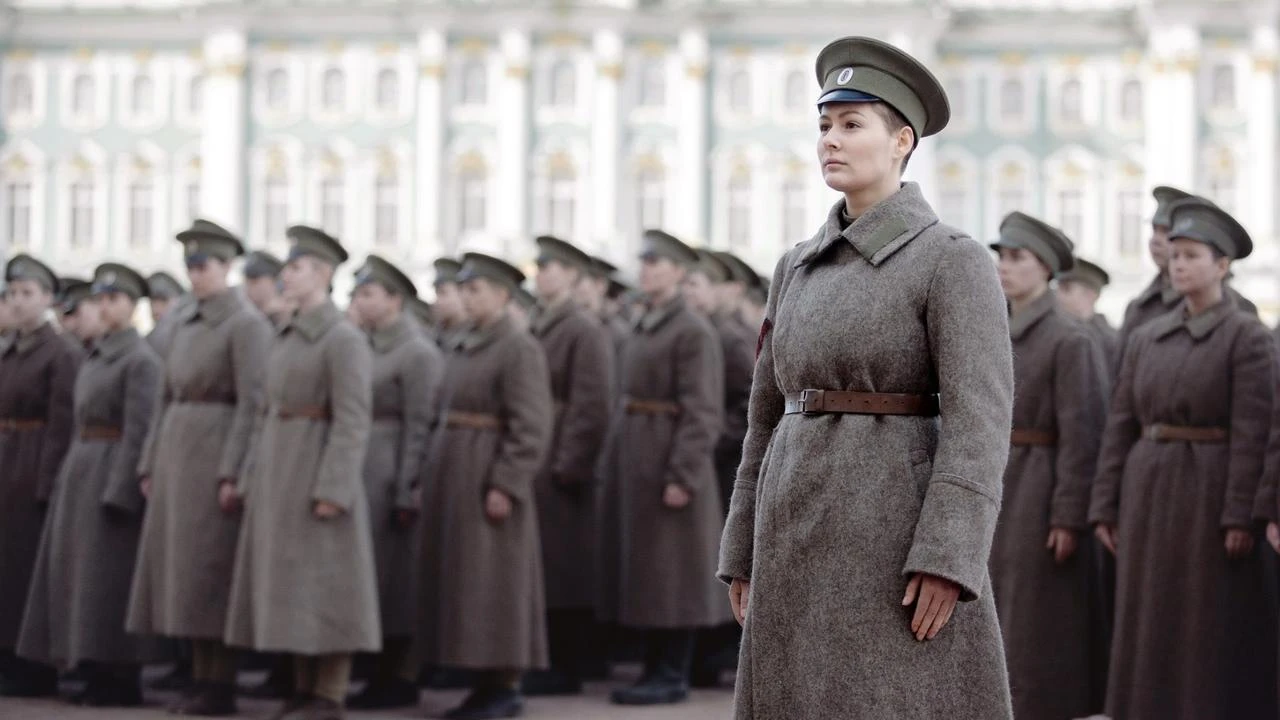 Fotograma de "Batallón" en el que aparece una mujer vestida de militar con un abrigo gris y una gorra. Detrás, hay todo un ejército de personas vestidas como ella.