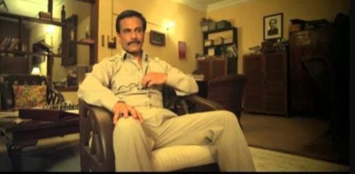 Un hombre con uniforme claro está sentado en una silla dentro de una habitación cálidamente iluminada. El entorno está decorado con libros, muebles de madera y una mesa con un tablero de ajedrez. Su expresión es seria.