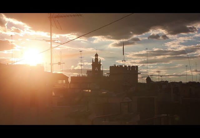 Atardecer, con algunas nubes, en una ciudad. El sol da de cara, dificultando la visión de los edificios.