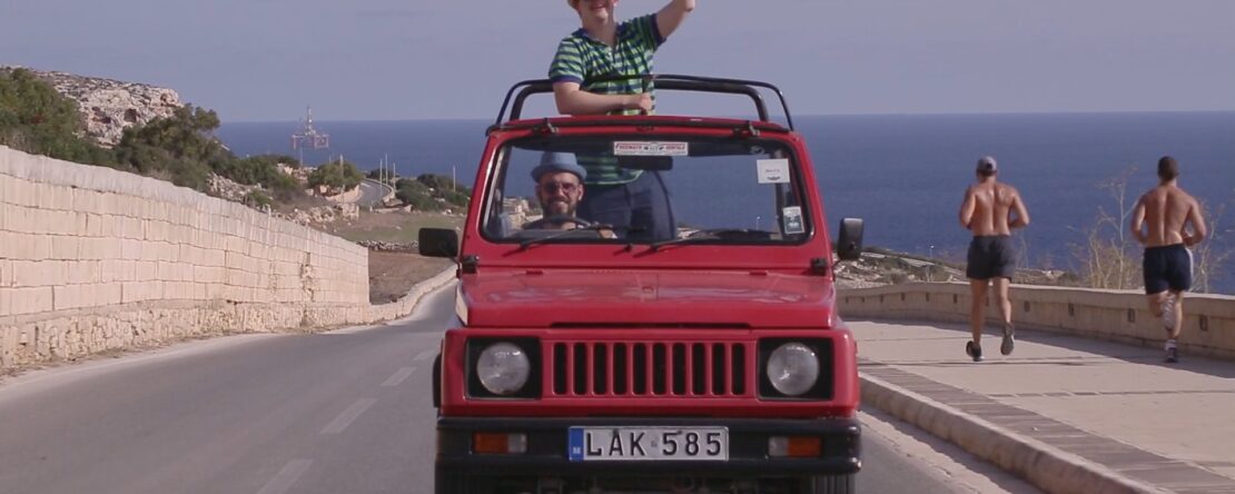 Fotograma de Altrove - En otra parte. La imagen muestra un coche rojo descapotable conduciendo por una carretera costera con el mar al fondo. El protagonista está de pie en el asiento del copiloto.
