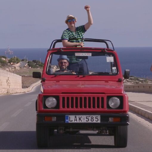 Fotograma de Altrove - En otra parte. La imagen muestra un coche rojo descapotable conduciendo por una carretera costera con el mar al fondo. El protagonista está de pie en el asiento del copiloto.