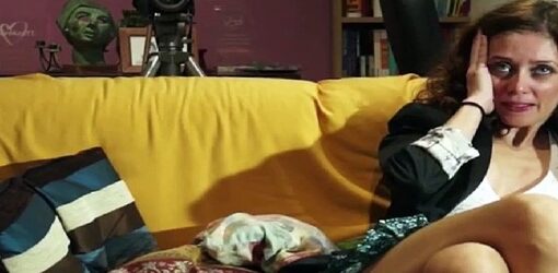 Una mujer con expresión angustiada esta sentada en un sofá amarillo en una sala colorida y desordenada. Frente a ella, un control remoto y un tablero de ajedrez sobre una mesa de cristal.