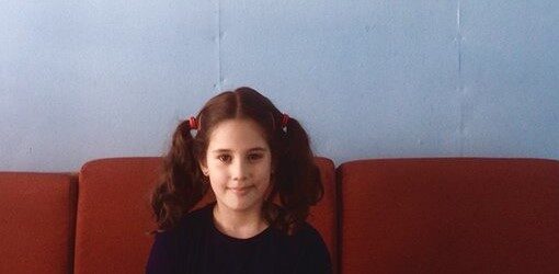 Una niña sonriendo en primer plano, sentada en un sofá rojo, sobre una pared azul.