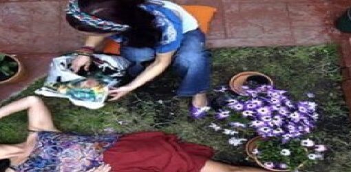 Una mujer yace en el suelo entre flores como si estuviera desmayada, con una pose dramática. A su lado, otra persona sigue jardineando tranquilamente.