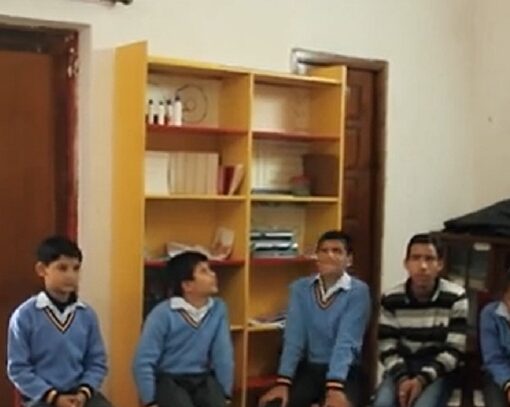 Un hombre de pie frente a un grupo de ocho niños sentados en sillas formando un semicírculo dentro de un aula. La mayoría lleva uniforme azul.
