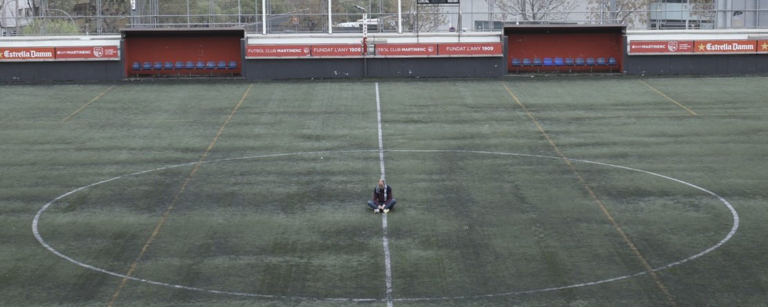 Imagen de un campo de futbol, en el centro del campo hay tres personas de pie.