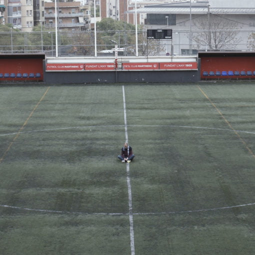 Imagen de un campo de futbol, en el centro del campo hay tres personas de pie.