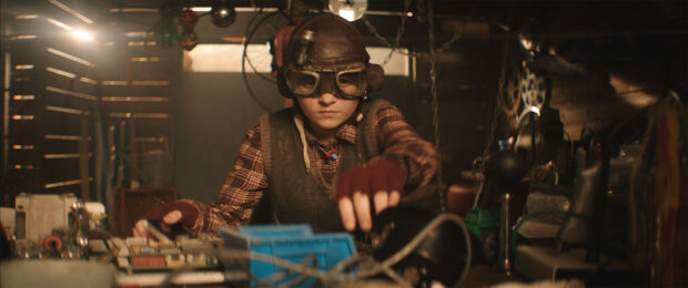 El fotograma de la película muestra a una de las protagonistas trabajando en un taller con herramientas y equipos esparcidos por el espacio. Lleva guantes sin dedos y viste una camisa de cuadros con chaleco. Estética steampunk.