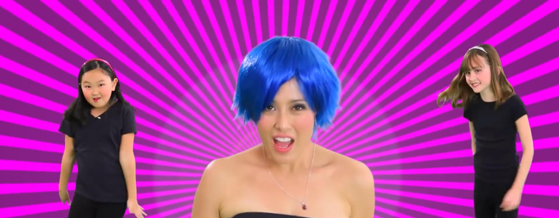 Fotograma de "Poesía y Melodía". Dos niñas a los lados y una mujer con peluca azul en el centro, sobre fondo morado, cantan y bailan