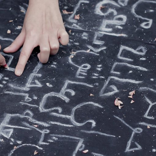 Una mano sostiene un bolígrafo y escribe símbolos extraños sobre un papel. Los trazos parecen abstractos o pertenecientes a un idioma desconocido.
