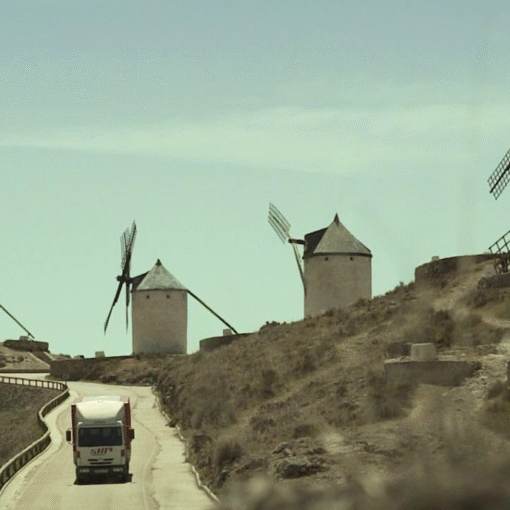 Un vehículo avanza por una carretera rural rodeada de molinos de viento tradicionales sobre una colina árida, bajo un cielo despejado, evocando tránsito, soledad y desplazamiento.