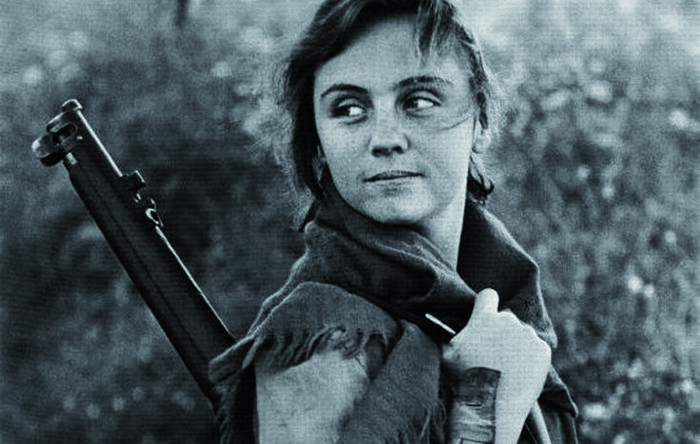 Fotograma de Cunetas en blanco y negro. Niña con ropa de campesina y un fusil en el hombro en el campo. Postura girada, mirando hacia atrás con media sonrisa, cargada de reivindicación y esperanza.