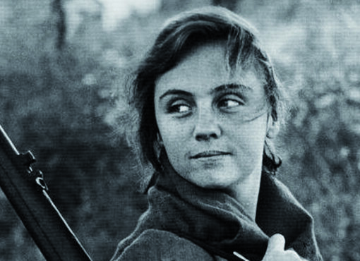 Fotograma de Cunetas en blanco y negro. Niña con ropa de campesina y un fusil en el hombro en el campo. Postura girada, mirando hacia atrás con media sonrisa, cargada de reivindicación y esperanza.