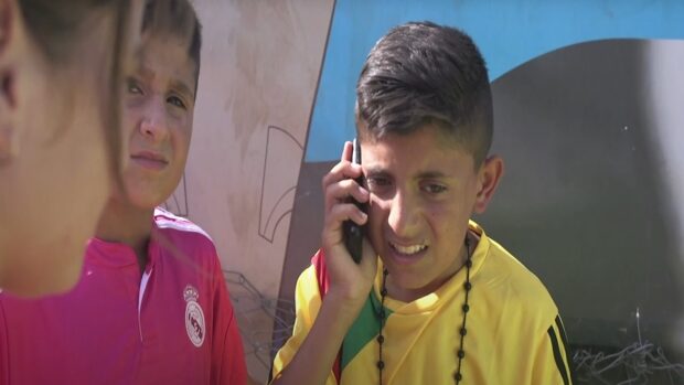 Muestra a dos jóvenes, uno con una camiseta roja de fútbol y el otro con una camiseta amarilla, hablando por un teléfono móvil.