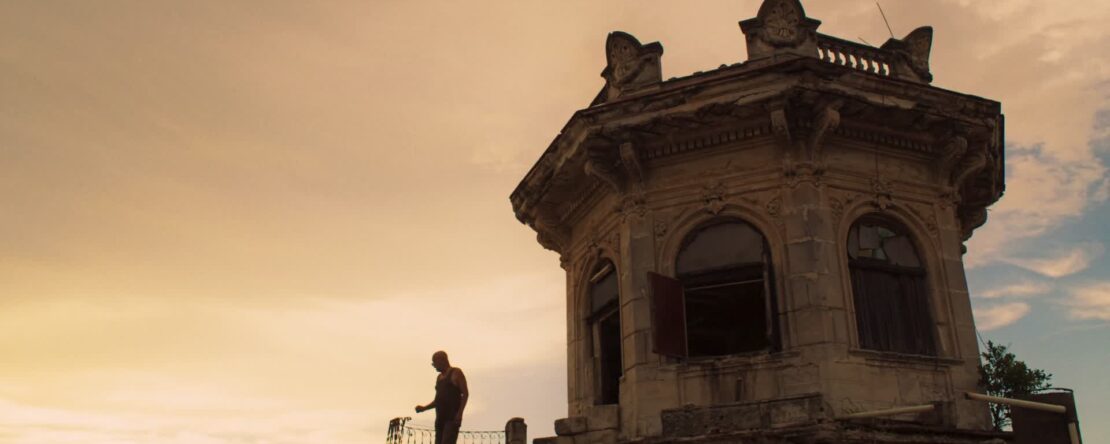 Fotograma de Arriba Habana en el que aparece un hombre subido a una torre muy alta. Al fondo, está el cielo con los colores anaranjados del atardecer.