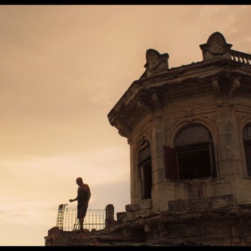 Fotograma de Arriba Habana en el que aparece un hombre subido a una torre muy alta. Al fondo, está el cielo con los colores anaranjados del atardecer.