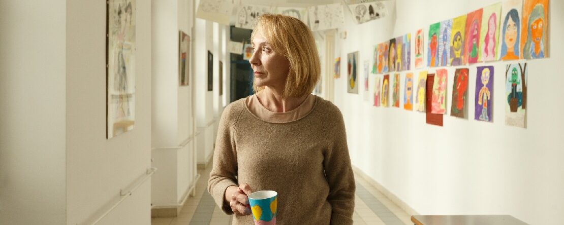 Una mujer rubia con suéter beige sostiene una taza colorida mientras camina por un pasillo decorado con dibujos infantiles.
